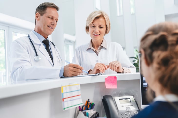 Two-doctors-colleagues-standing-at-reception-desk-talking-and-sharing-information-with-receptionist-in-modern-clinic-1275072738_3867x2579-3